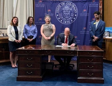 Individuals stand around the Governor as he signs a bill with the Colorado State Seal in the background.