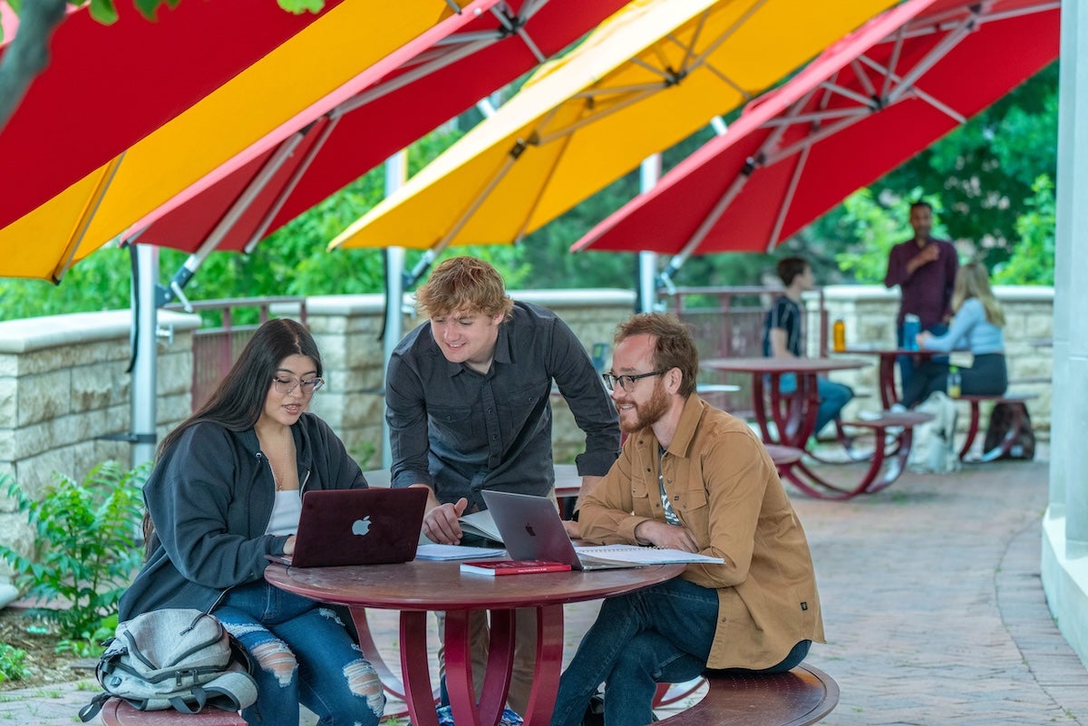 3 students look at a laptop on a picnic table under red and yellow umbrellas on the University of Denver campus. 