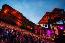 Photo of Red Rocks Amphitheatre at night during a concert with pink lighting on the stage, a large red rock visible in the background and crowded seats in the foreground