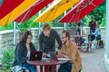 3 students look at a laptop on a picnic table under red and yellow umbrellas on the University of Denver campus. 
