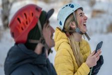 Two students smile as they wear climbing helmets and look upwards while one holds a cellphone. 