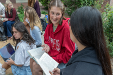 A student in a University of Denver sweatshirt holds open a book while talking with another student on the University of Denver campus. 