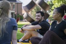 A group of three students sits and chats on the grass on campus.