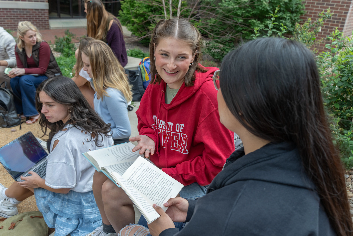 A student in a University of Denver sweatshirt holds open a book while talking with another student on the University of Denver campus. 