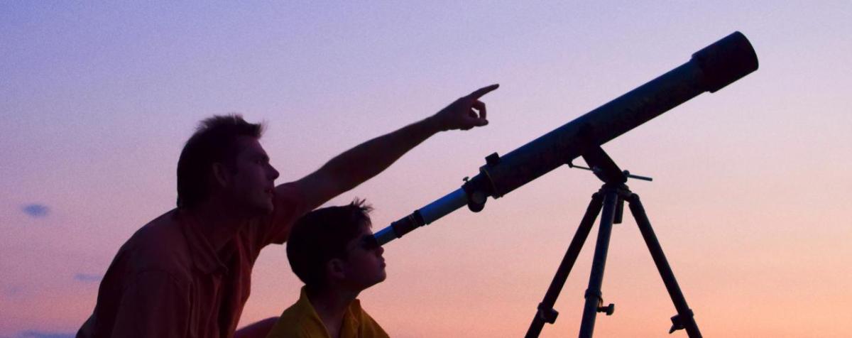 An adult and child look through telescopes during a sunset. The adult is pointing towards the sky.