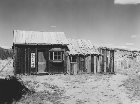 A black and white image of a wooden cabin with a sign in Chinese to the left of of the front door.