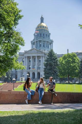 Three students sit in front of the gold dome of the Colorado State Capitol. 