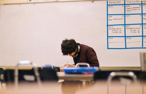 A student with headphones around his neck works in a classroom by himself. 