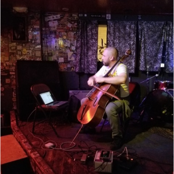 Kevin plays a cello on a stage in a cozy music venue. 