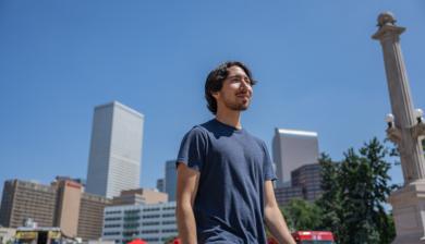 A student walks in downtown Denver with the city skyline behind him 