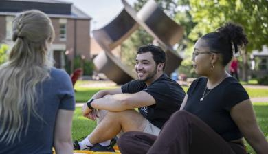 A group of three students sits and chats on the grass on campus.