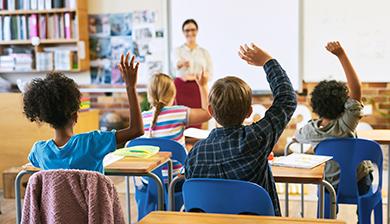 Seen from the back of a classroom, rows of young students raise their hands as their smiling teacher looks on.