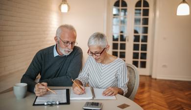 An elderly couple sits at a table in front of a computer and a calculator.