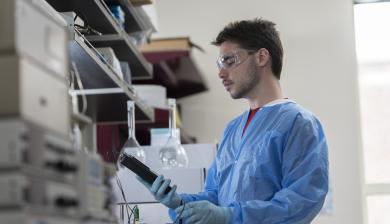 Image of a young man in blue lab scrubs looking at a small handheld machine in a lab.