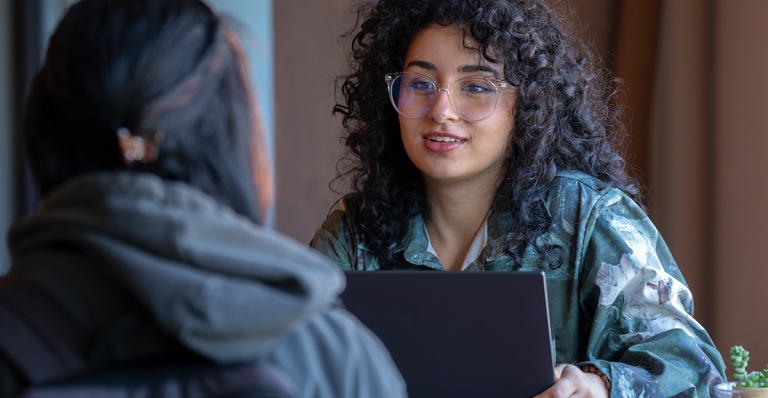 A student holds her laptop screen while listening to another student.