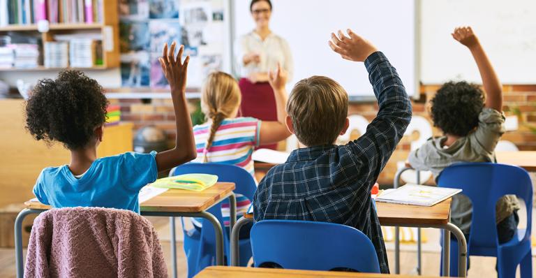 Students raise their hands at their desk to respond to the teacher in a classroom.